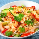 A vibrant Asian noodle salad served in a white bowl, featuring a colorful mix of fresh vegetables, herbs, and thin noodles, garnished with sesame seeds and a lime wedge, set against a rustic wooden background