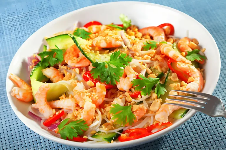 A vibrant Asian noodle salad served in a white bowl, featuring a colorful mix of fresh vegetables, herbs, and thin noodles, garnished with sesame seeds and a lime wedge, set against a rustic wooden background