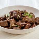 A close-up view of juicy garlic steak bites on a plate. The steak bites are golden brown and caramelized on the outside, with tender, medium-rare steak in the center. They are drizzled with a garlicky butter sauce and garnished with fresh parsley.