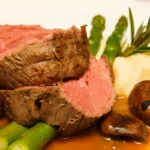 Sliced prime rib arranged on a serving platter, surrounded by roasted asparagus spears and golden brown mushrooms. Fresh herbs garnish the dish.